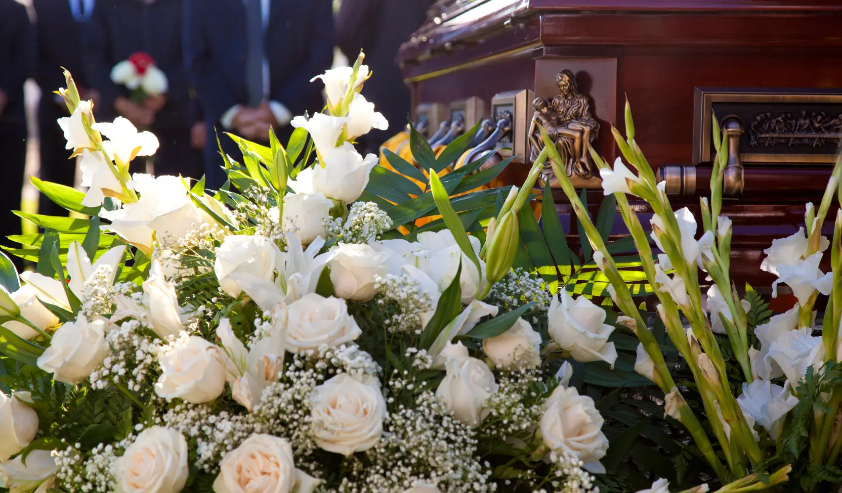 Dunkelroter Holzsarg mit Bronzebeschlägen und ein großes Gesteck aus weißen Rosen, Lilien und Schleierkraut, im Hintergrund Trauergäste