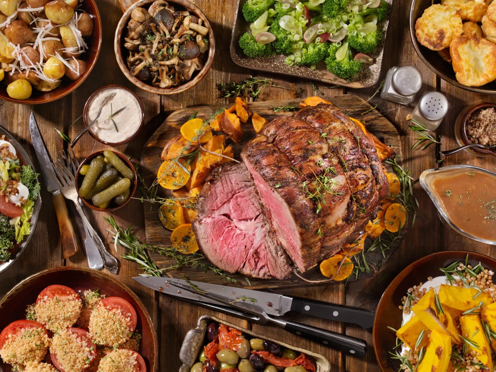 BBQ-Buffet auf dem Holztisch mit aufgeschnittenem Rinderbraten, Röstkartoffeln, Pilzen, Brokkoli, Salat, Oliven und Bratensauce