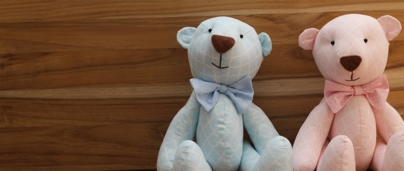 Zwei Stoff-Teddybären in Blau und Rosa vor einer warmen Holzwand